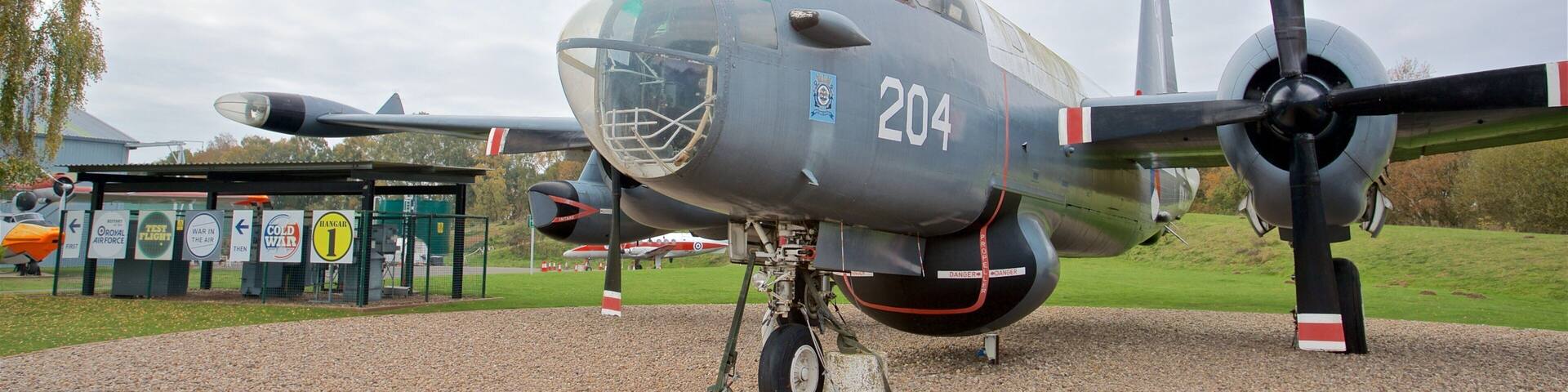 A military aircraft with the number 204 displayed on its nose, parked on a gravel surface with a grassy field and trees in the background.