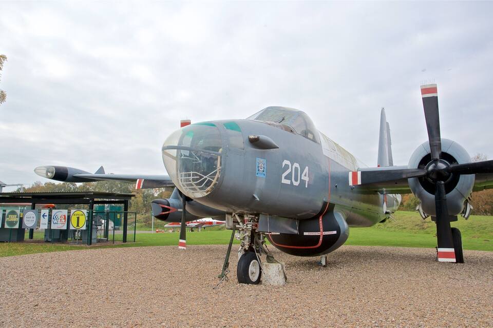 Un aereo militare con il numero 204 sullo scafo, parcheggiato su un terreno ghiaioso, con un campo erboso e alberi sullo sfondo.
