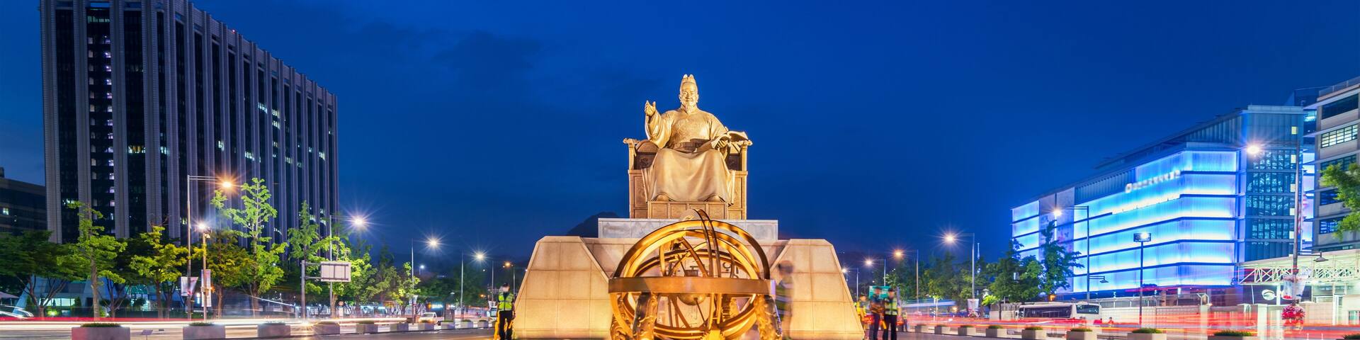 Una vista nocturna de la ciudad con una estatua destacada en el centro, rodeada de edificios modernos y una amplia plaza.