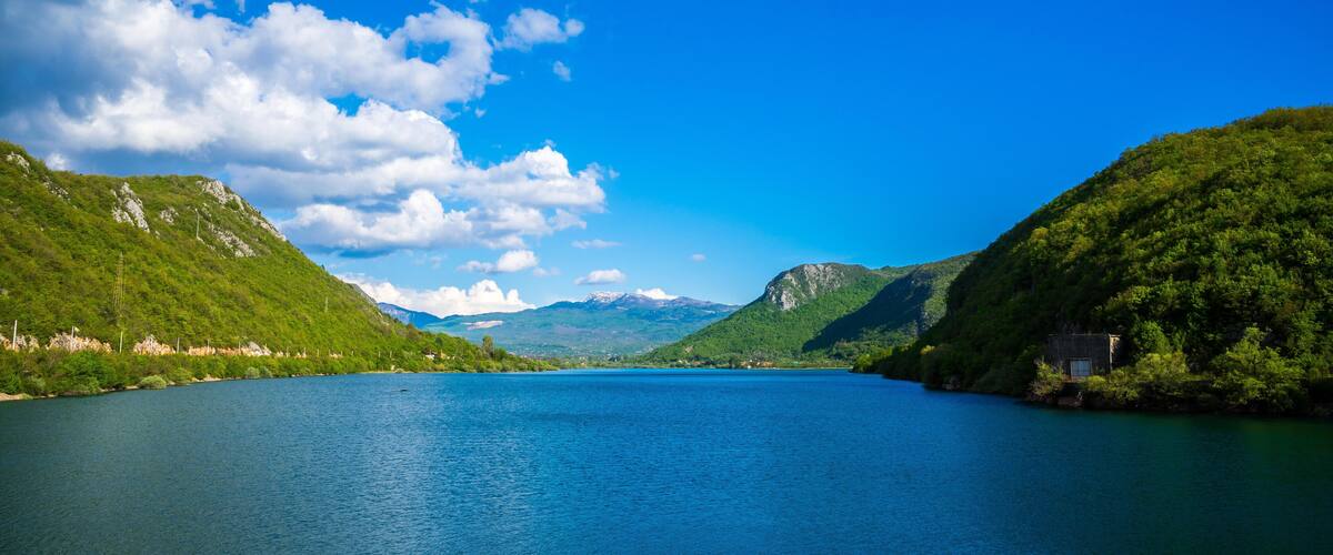 A calm lake surrounded by lush green hills and mountains under a clear blue sky.