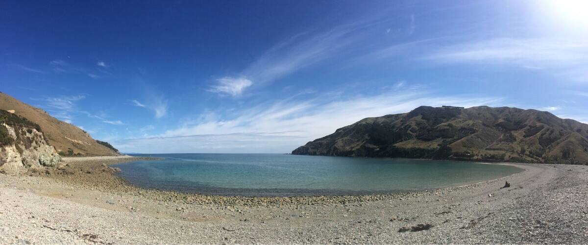 A pebble beach with a clear blue sea, rocky hills, and a distant mountain.