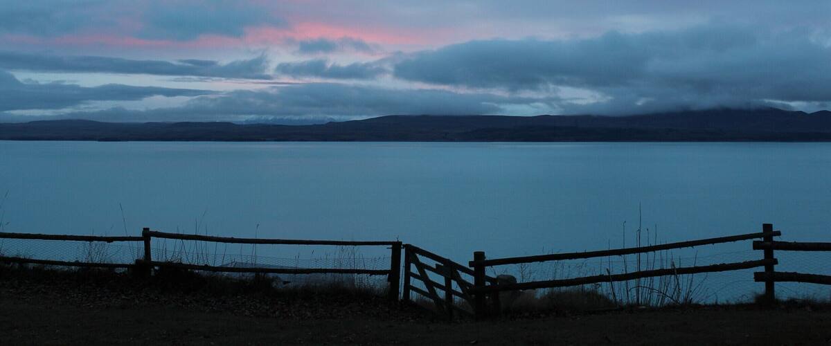 A lake with a wooden fence in the foreground and mountains in the background under a cloudy sky.