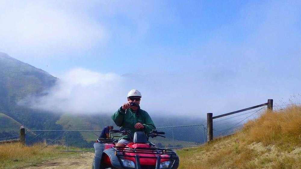 Una persona conduce un vehículo todo terreno rojo por un sendero de tierra, con montañas y niebla al fondo.