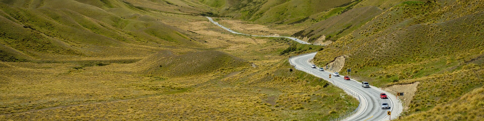 Uma estrada sinuosa nas montanhas, com veículos, cercada por colinas ondulantes e um céu nublado.