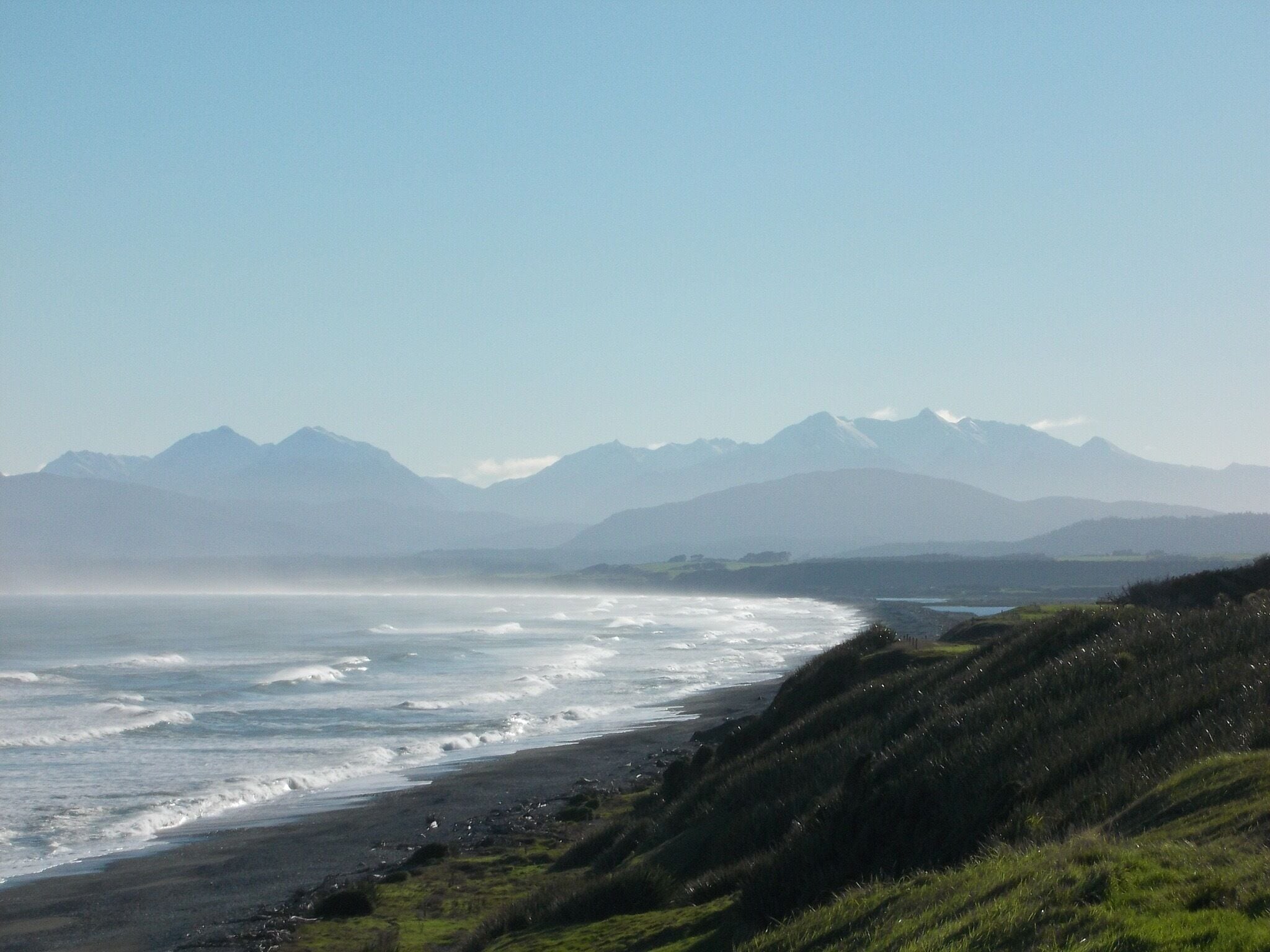 A coastal landscape with a beach, waves, and distant mountains.