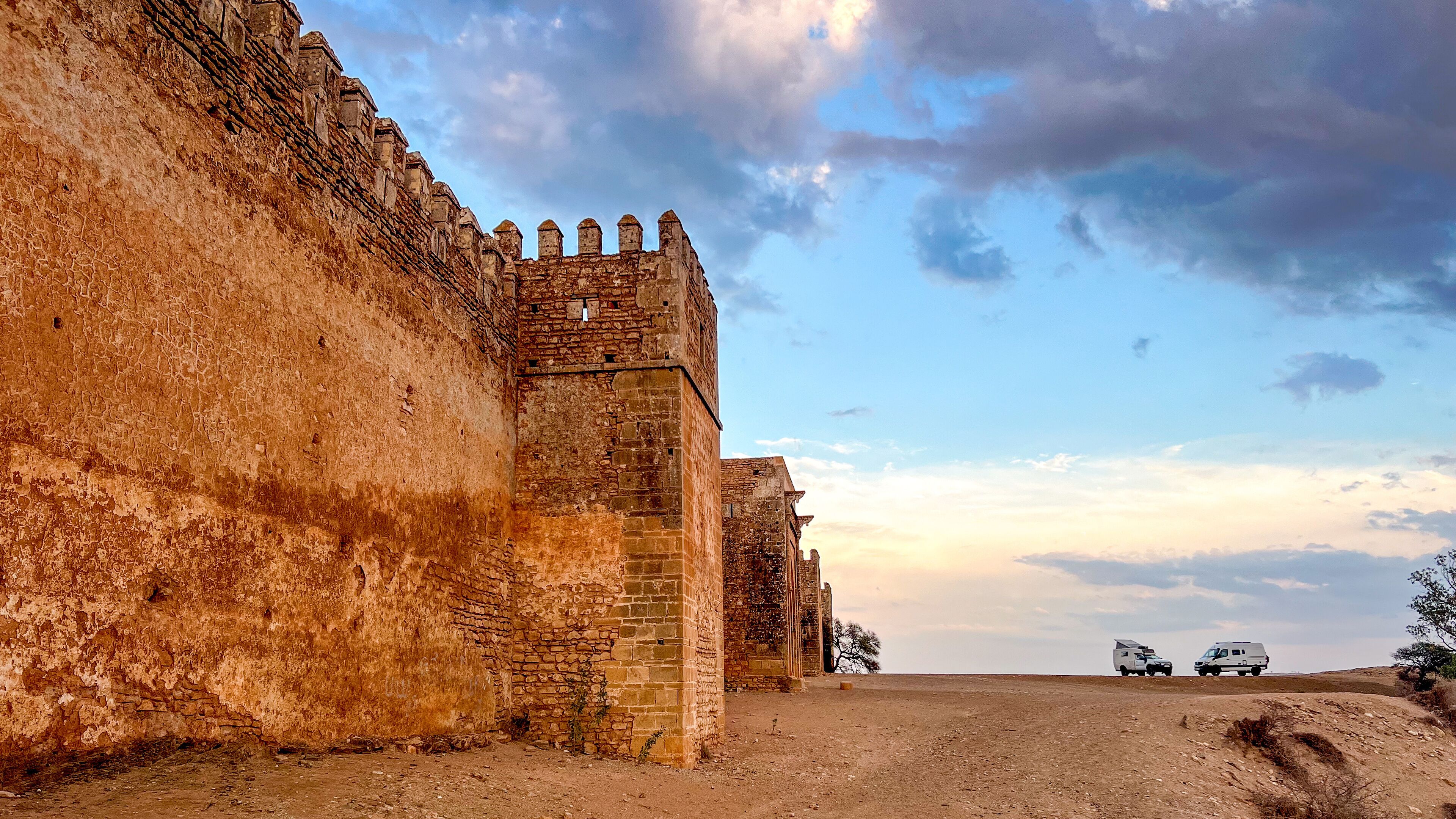 A historic stone wall with battlements, a clear sky, and a dirt road with vehicles.
