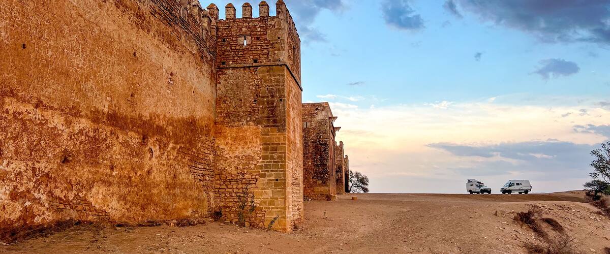 A historic stone wall with battlements, a clear sky, and a dirt road with vehicles.