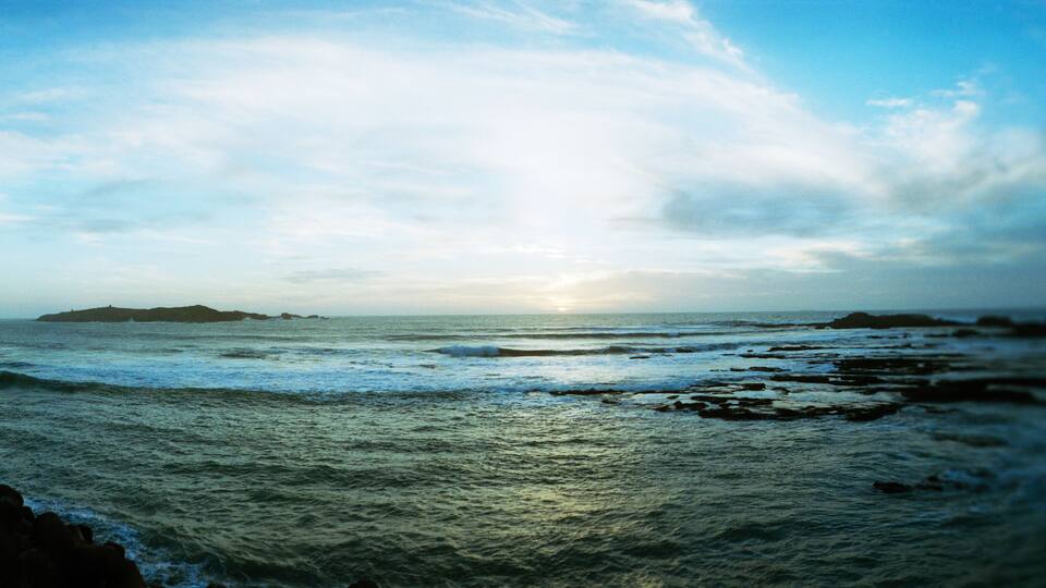 A coastal landscape with waves crashing against rocks and a distant island under a cloudy sky.