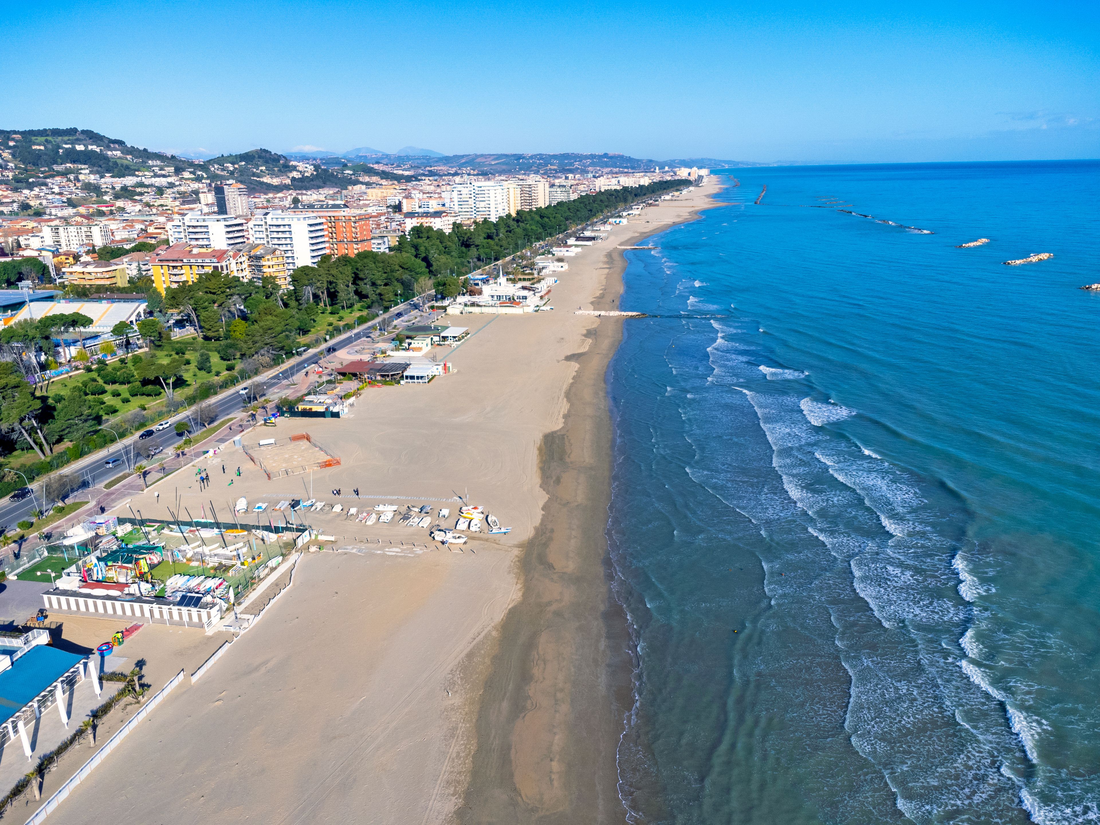 Un paisaje urbano costero con una extensa playa de arena, un mar azul cristalino y una zona urbana densa con edificios y zonas verdes.