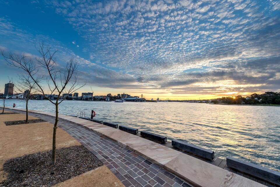 A riverside walkway with trees, a bridge, and a city skyline at sunset.
