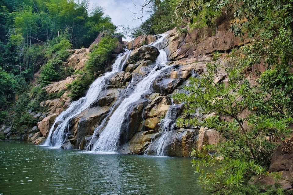 Una cascada en varios niveles que se precipita sobre un río tranquilo, rodeado de vegetación densa y terreno rocoso.