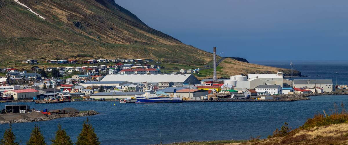 A coastal town with a large industrial facility, a harbor with boats, and a mountainous backdrop.