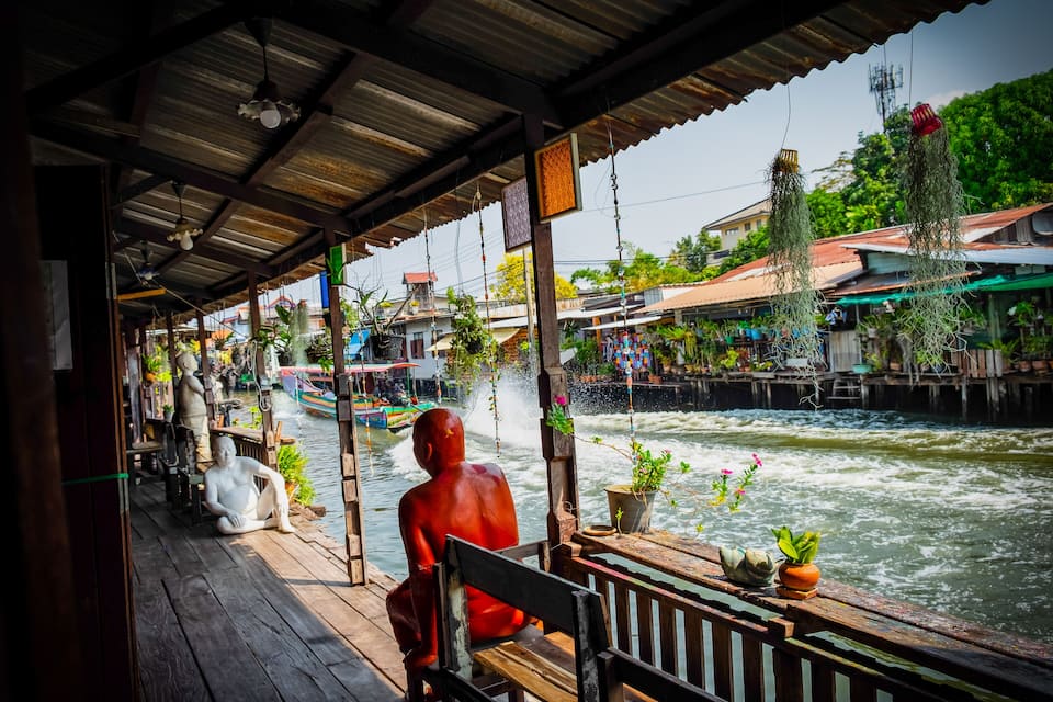 A wooden dock with a red statue, potted plants, and a river with boats.