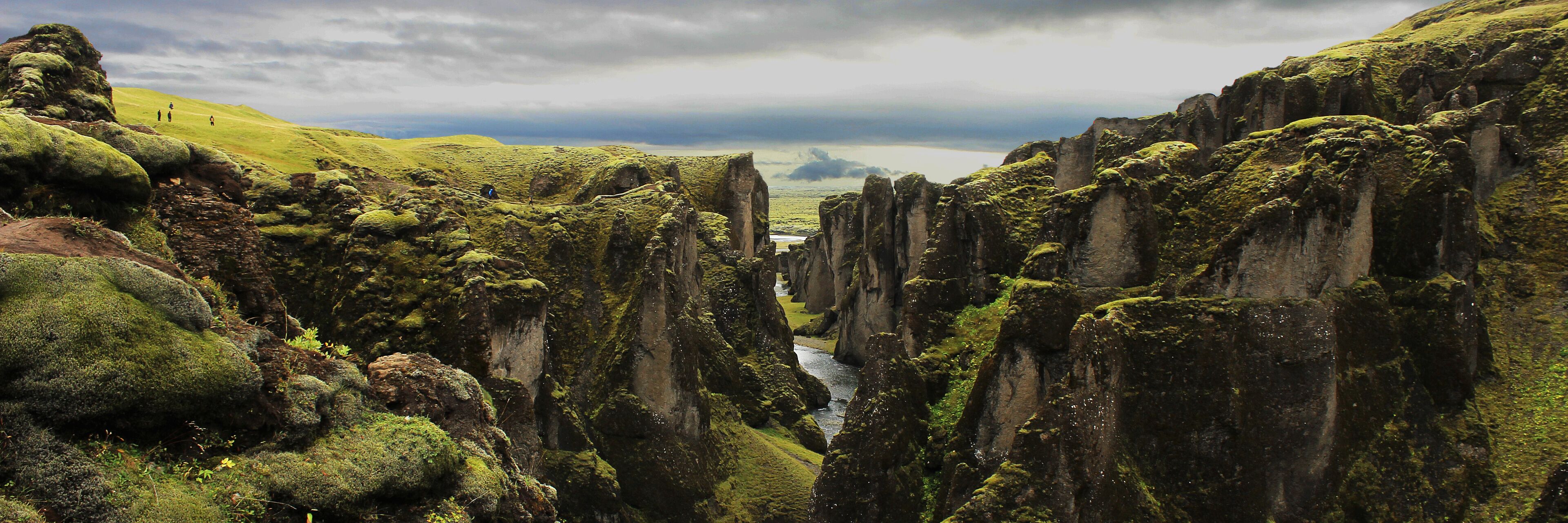 A narrow canyon with tall, moss-covered rock formations and a distant view of a body of water under a cloudy sky.