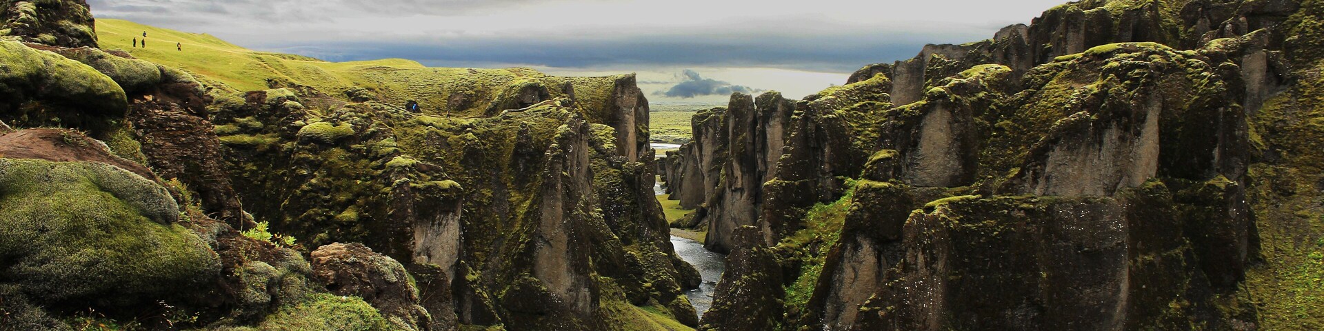 A narrow canyon with tall, moss-covered rock formations and a distant view of a body of water under a cloudy sky.