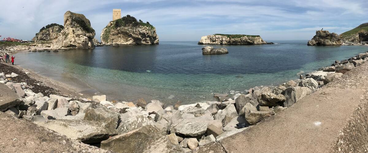 A coastal landscape with rocky formations, clear waters, and a tower on a prominent rock.