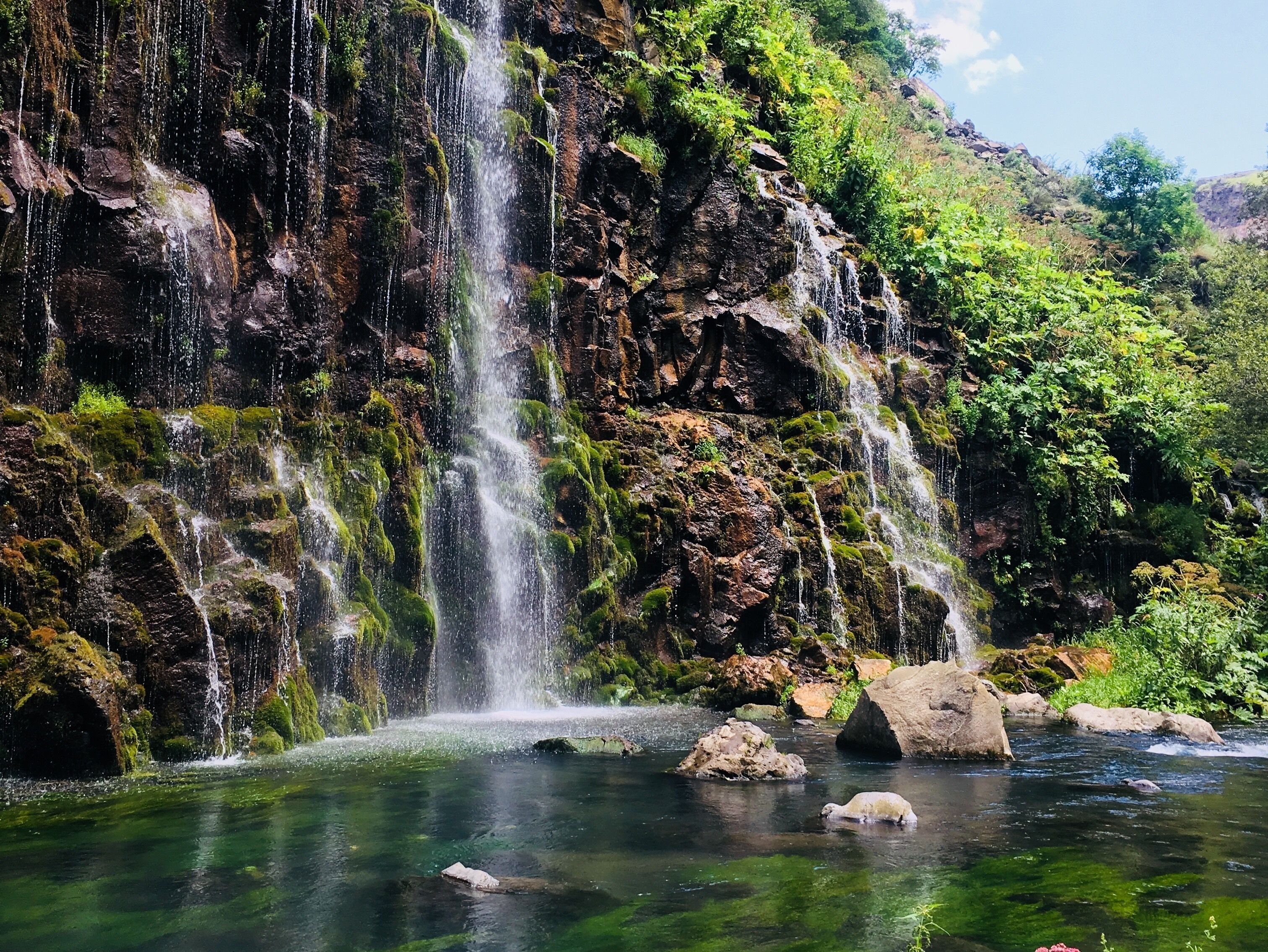 A waterfall cascading into a clear river surrounded by rocky terrain and lush greenery.