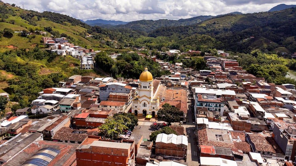 A town with a prominent church featuring a golden dome, surrounded by densely packed buildings and lush green hills in the background.