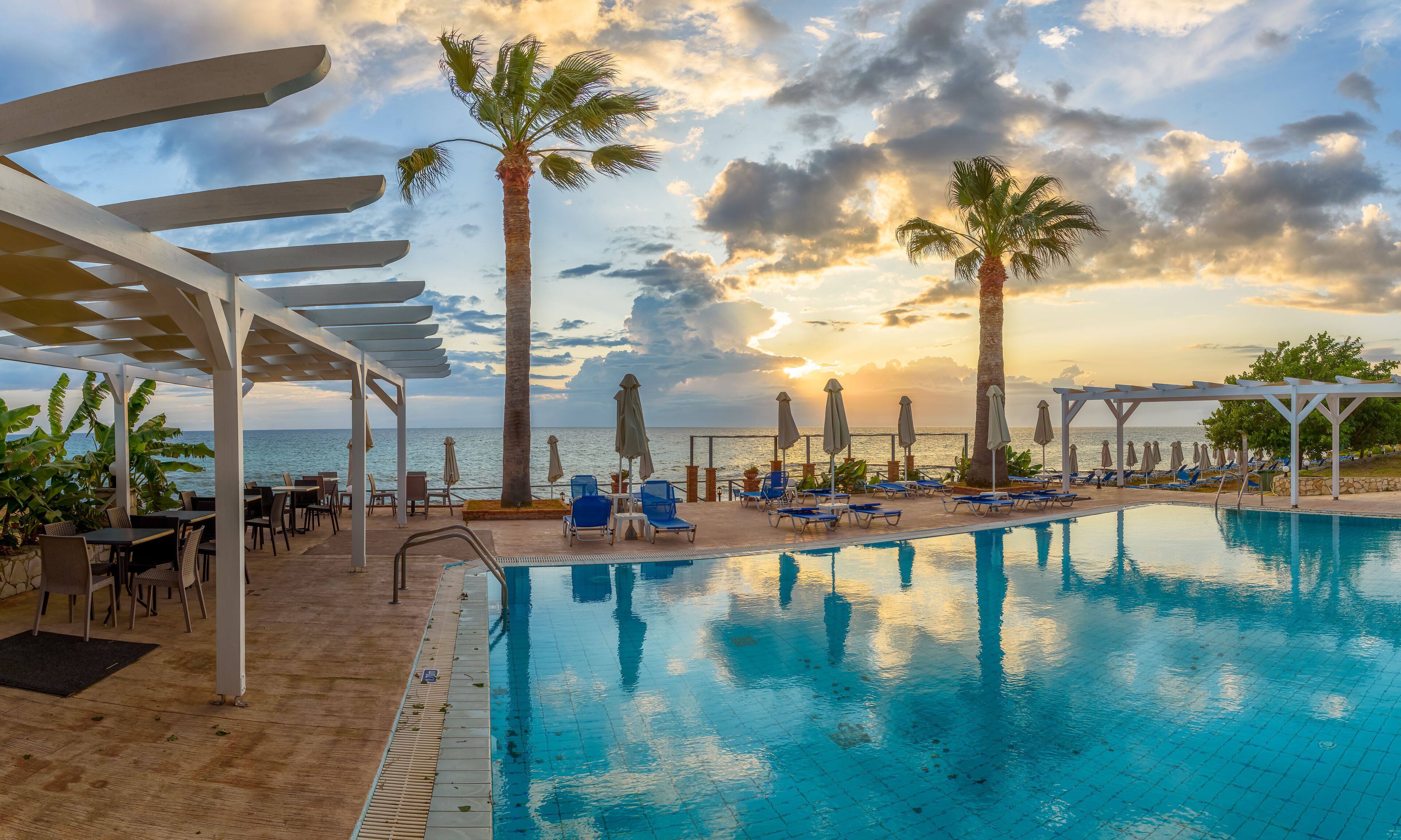 A poolside area with lounge chairs, palm trees, and a view of the ocean at sunset.