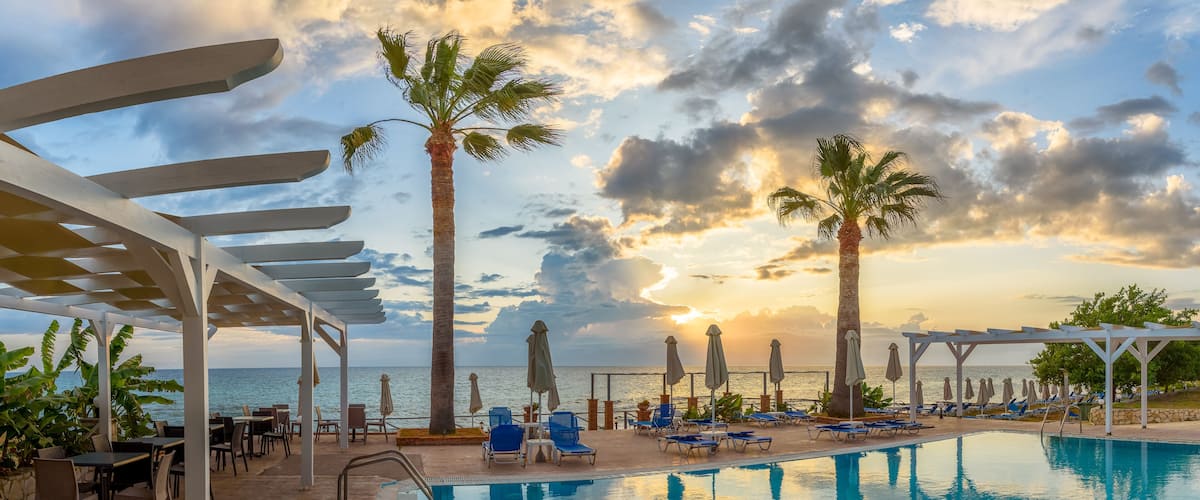 A poolside area with lounge chairs, palm trees, and a view of the ocean at sunset.