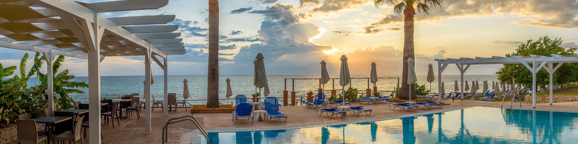 A poolside area with lounge chairs, palm trees, and a view of the ocean at sunset.