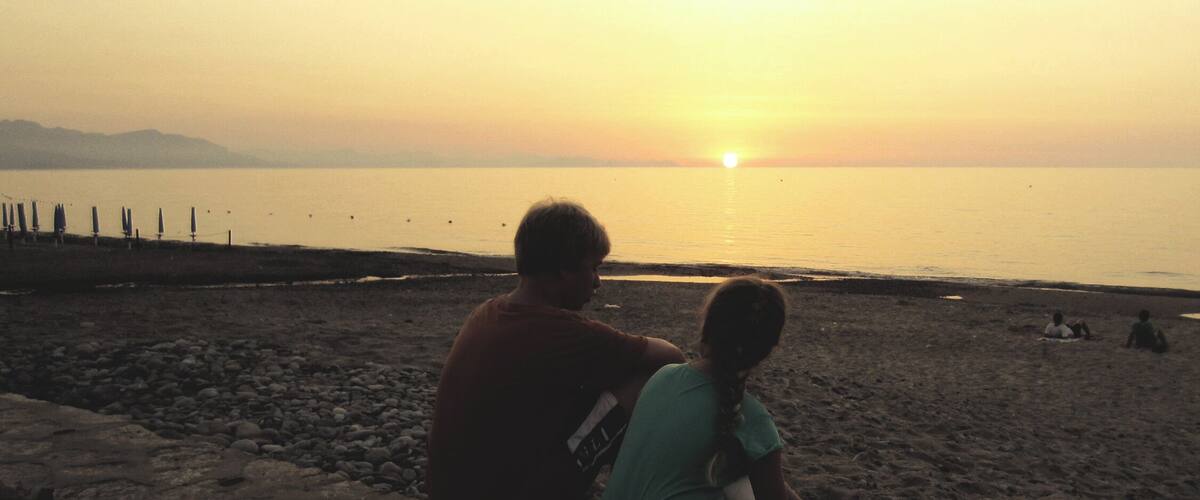 A sunset over a calm sea with a rocky shoreline and a few people on the beach.