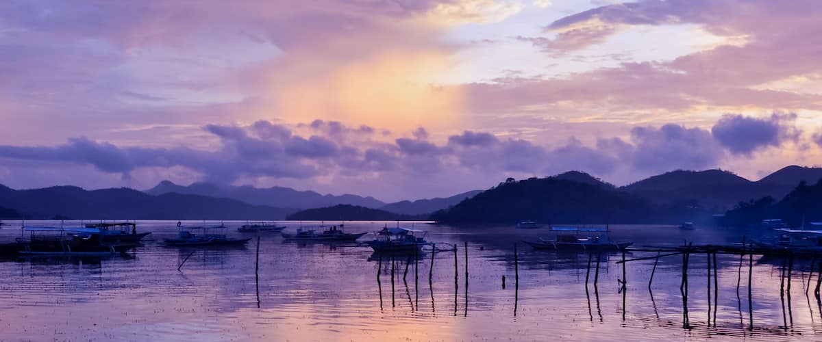 A calm lake with boats and a mountainous landscape during sunset.