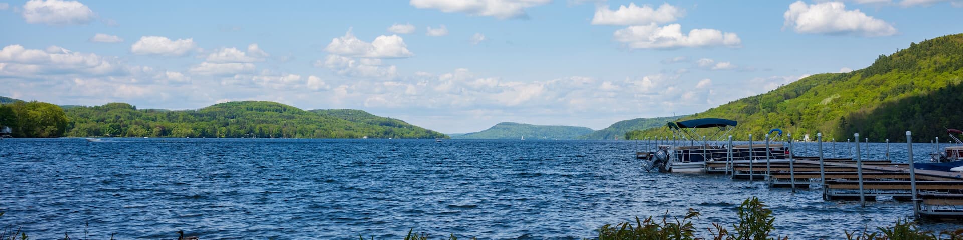 The blue waters of Otsego Lake in Cooperstown, New York, on a sunny summer day with cumulus clouds in the sky, photographed near a dock.