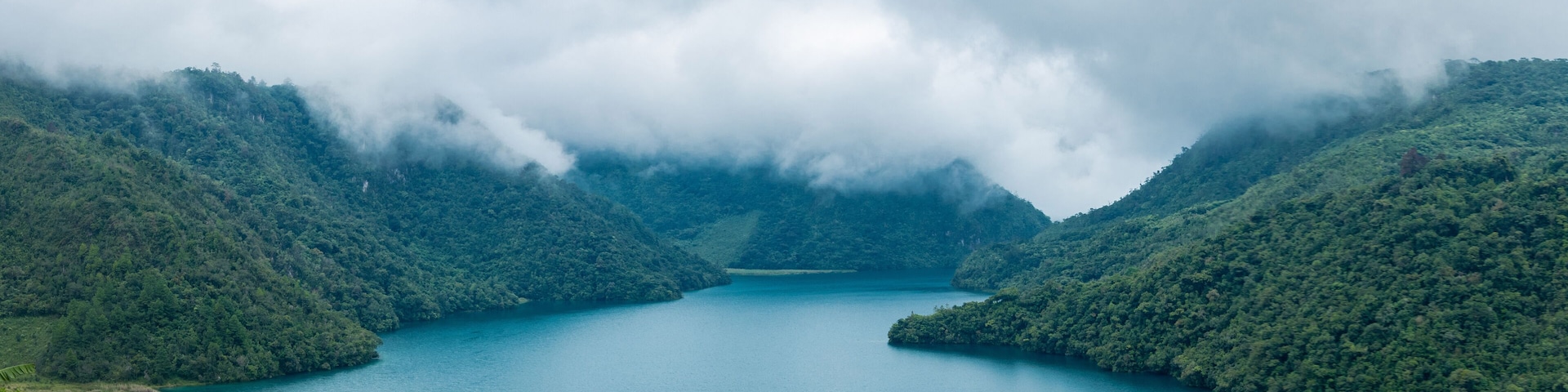 Un lago rodeado de colinas cubiertas de exuberante vegetación y un bosque denso, parcialmente oculto por nubes.