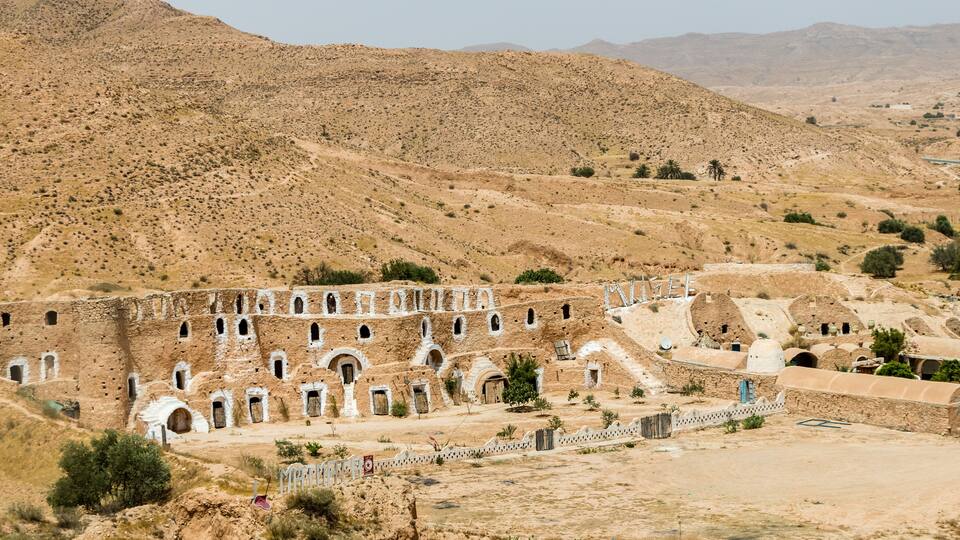 A historical site with ancient architecture, including a large building with multiple arched doorways and a fortress-like structure.