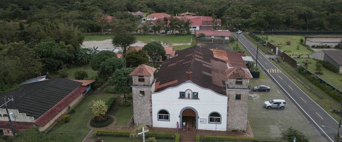 A white church with a bell tower, surrounded by lush greenery and a mountain in the background.