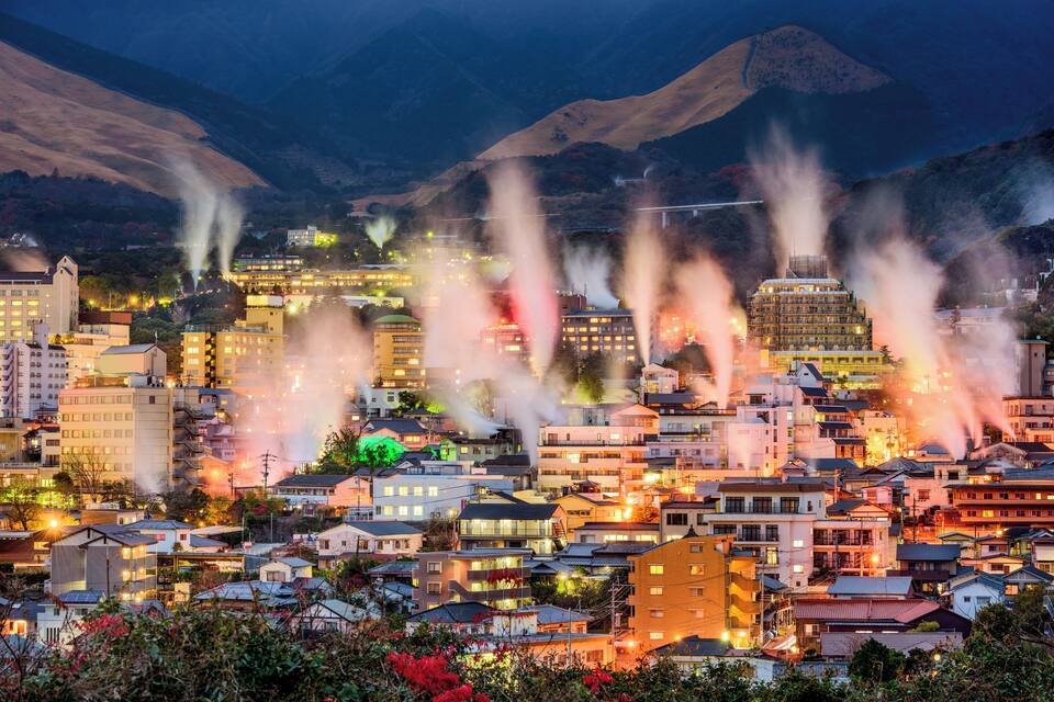 A cityscape with buildings and smoke plumes against a mountainous backdrop.