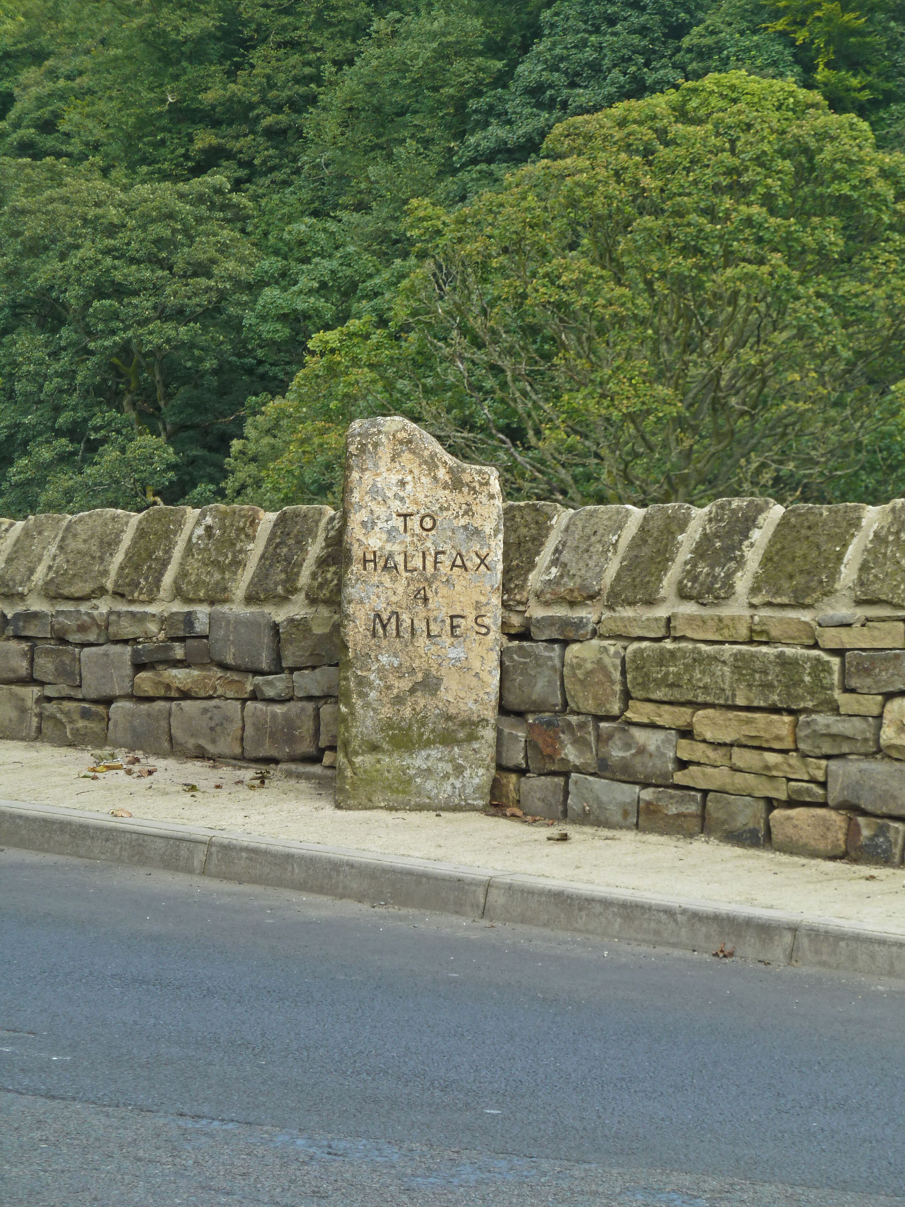 A stone marker with "TO HALIFAX 4 MILES" engraved on it, set against a backdrop of a stone wall and lush greenery.