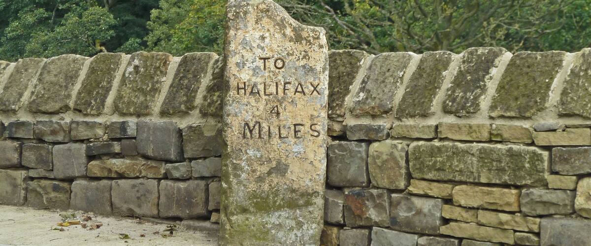 A stone marker with "TO HALIFAX 4 MILES" engraved on it, set against a backdrop of a stone wall and lush greenery.