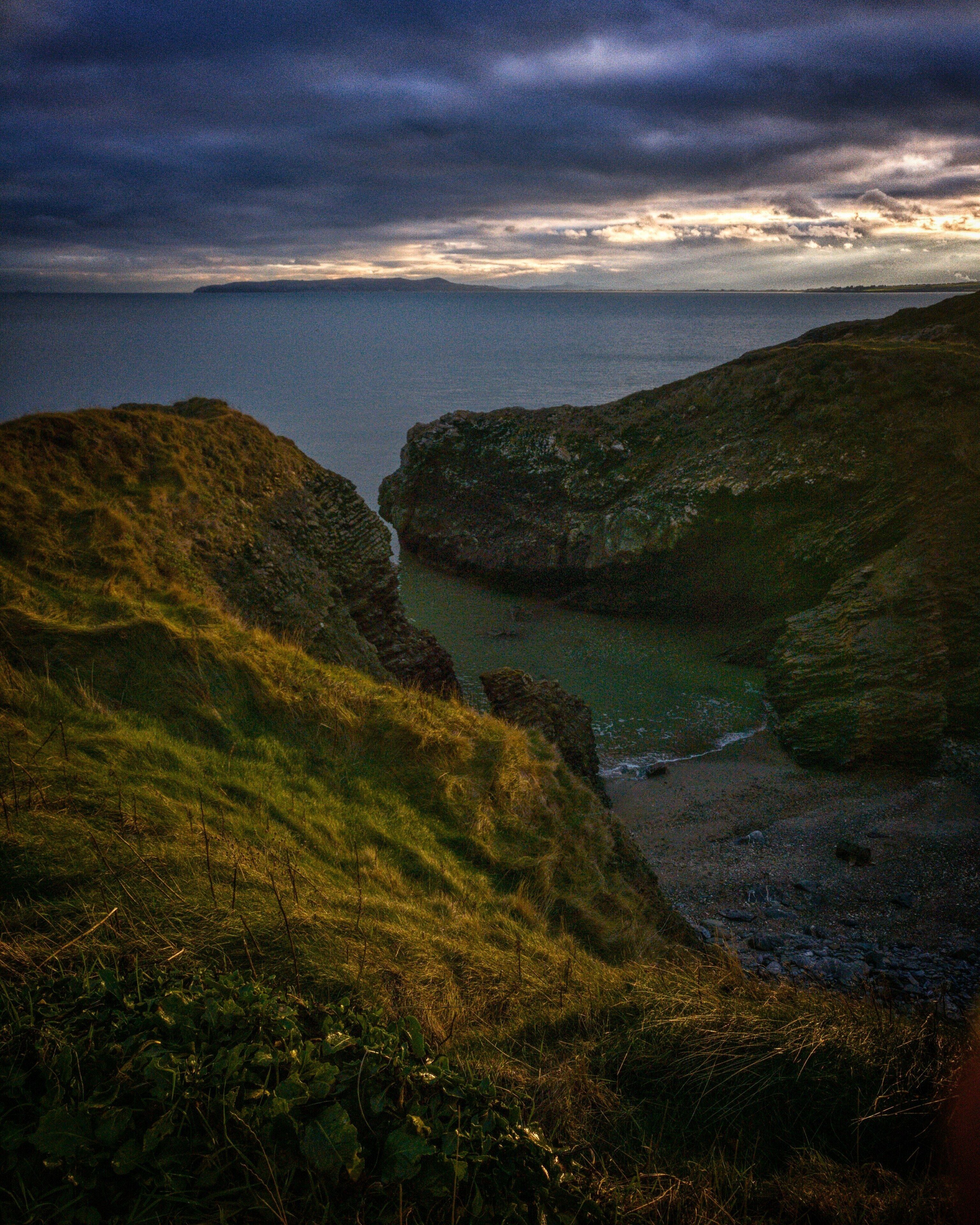 A coastal landscape with grassy hills and a rocky shoreline.
