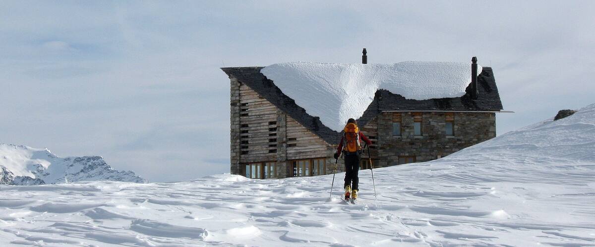 A person skiing towards a stone house on a snowy mountain.