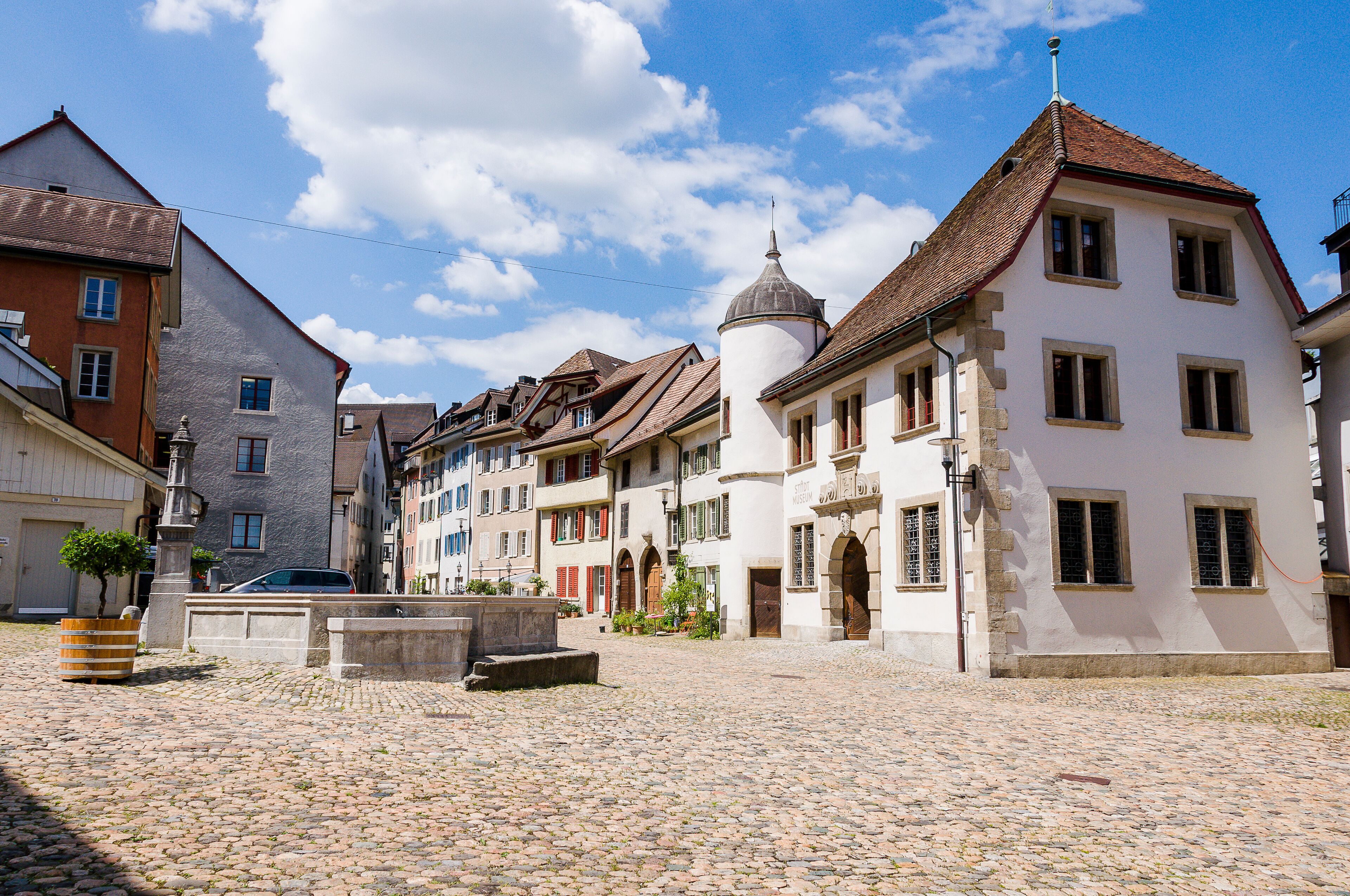 A cobblestone street lined with historic buildings, a fountain, and a clear blue sky.