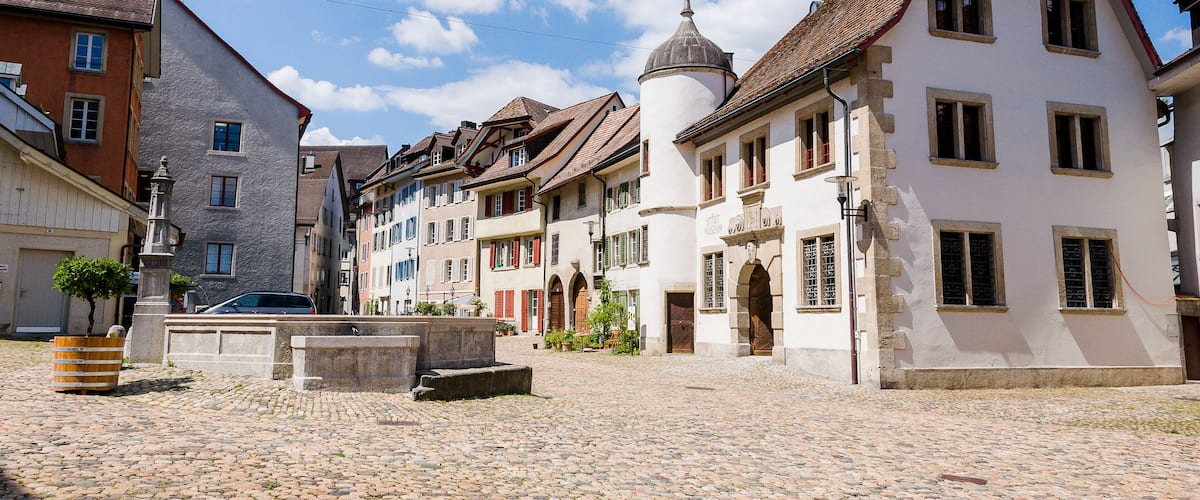 A cobblestone street lined with historic buildings, a fountain, and a clear blue sky.