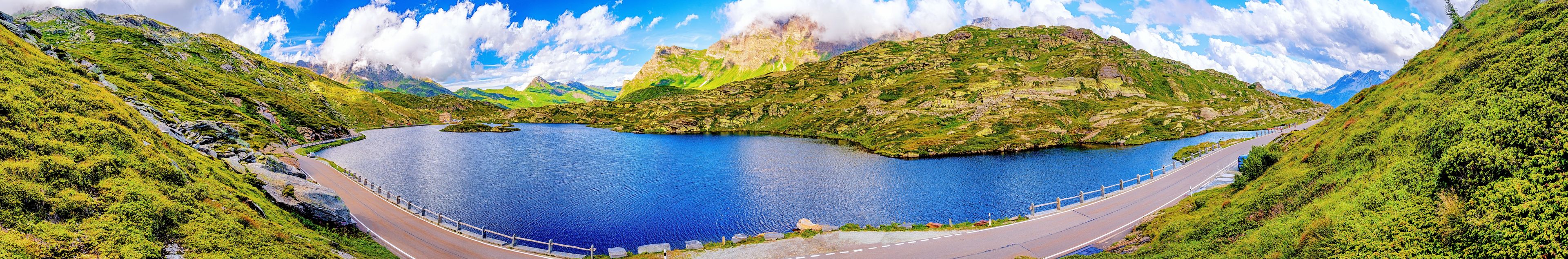 Une route sinueuse longe un grand lac, bordée de collines verdoyantes et de montagnes.