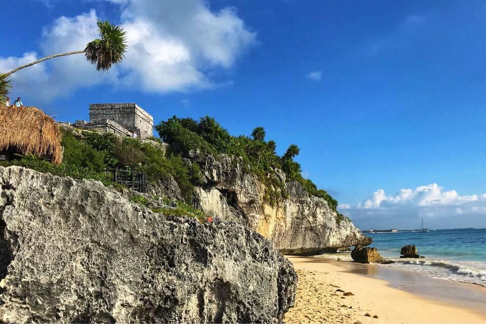 Paisagem costeira com uma estrutura de pedra em um penhasco, palmeiras e uma praia de areia.
