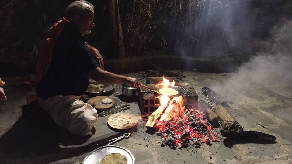 A person cooking outdoors with a fire, cooking utensils, and food on the ground.