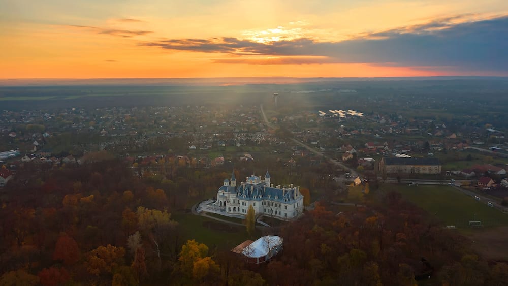 Eine Vogelperspektive eines historischen Schlosses, umgeben von Herbstlaub, mit einer Stadtansicht im Hintergrund.