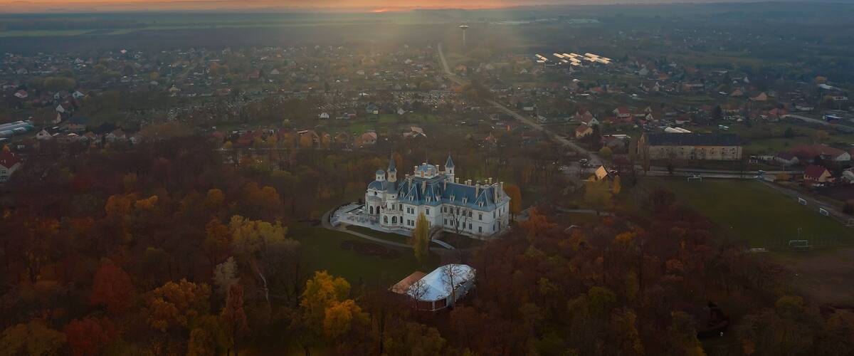 An aerial view of a historic castle surrounded by autumn foliage, with a cityscape in the background.