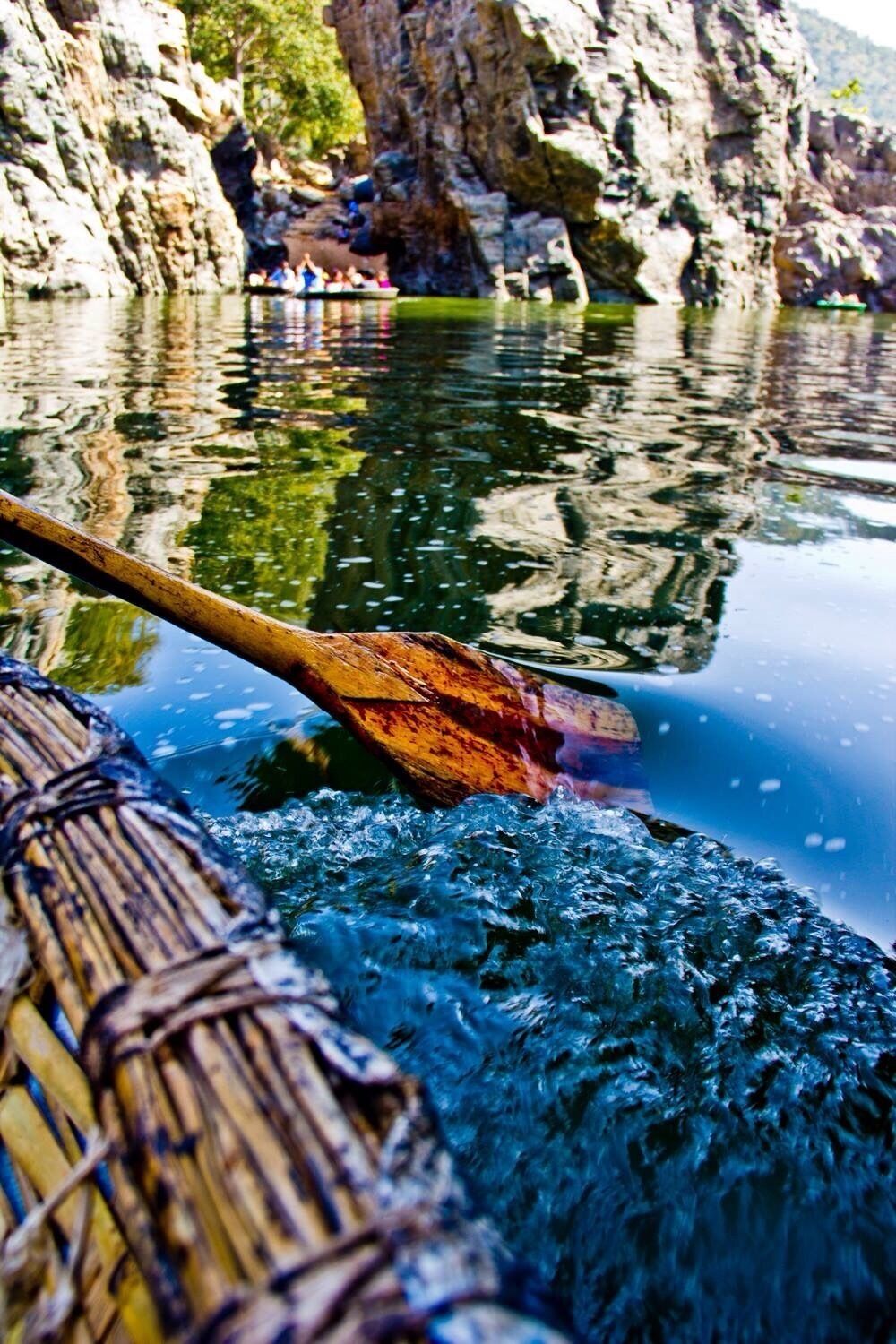 A wooden paddle in water, a rocky cliff, and a group of people in the background.