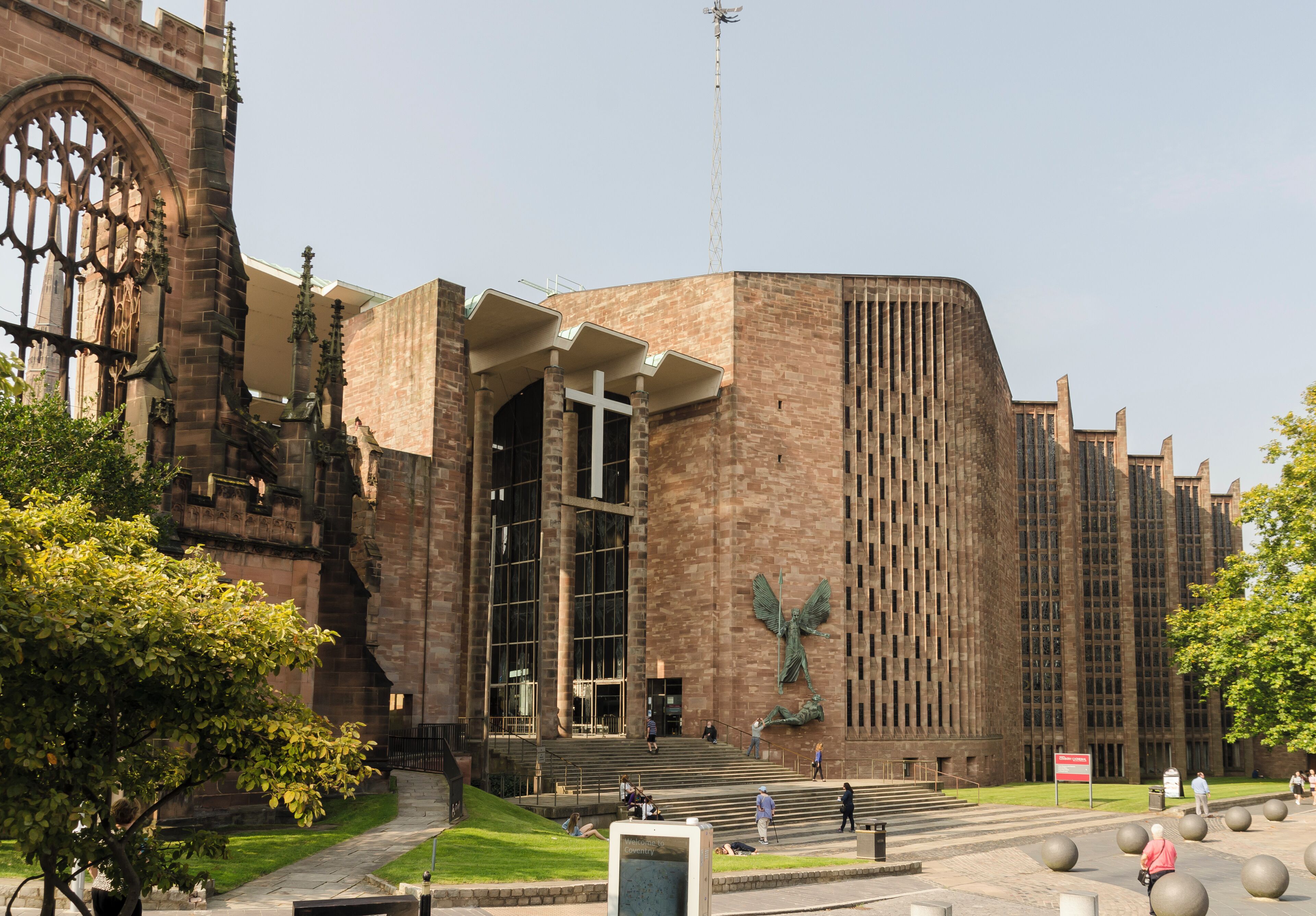 The Cathedral was built between 1956 and 1962 and was designed by Sir Basil Spence, following the destruction of the old mediaeval Cathedral in 1940 through enemy bombing. It was decided to build the new Cathedral adjacent to the old one, which was left in its semi-ruined state with its undamaged 14th century tower. It would in fact be the third Cathedral in Coventry, as this had been a religious site for nearly 1000 years. There was a competition for the design which was won by the Scottish architect Basil Spence. The unorthodox design was intended to be "a casket of jewels" and contains many works from famous artists. The Cathedral was consecrated on 25 May 1962. The Cathedral was built at right angles to the old one, and therefore aligns north-south. It is built in red Hollington sandstone with green slate cladding and a concrete roof. There is a nave of seven bays, although the aisles are full height, with slender piers tapering at the base. They rise to a concrete ribbed canopy with timber between the ribs. There is no clerestory. The nave walls are angled in a sawtooth fashion with tall stained-glass windows. The nave and chancel are as one. The (liturgical) west end has a fully glazed wall with engraved figures of saints and angels by John Hutton. The (liturgical) east wall has no window, but is dominated by a large tapestry of Christ in Glory by Graham Sutherland. There are two chapels, one to the north east (The Chapel of Christ the Servant) and one to the south west (The Chapel of Unity). The baptistery area to the south east has a full height slightly curved window with closely spaced mullions containing nearly 200 small windows. It was designed by John Piper and painted by Patrick Reyntiens. The font is made with a boulder from near Bethlehem. On the exterior wall is a sculpture by Epstein of Saint Michael and Lucifer. The low roof is crowned by a metal fleche topped by a cross. The organ is by Harrison and Harrison, divided in galleries either side of the altar. There is an open western (southern) porch linking the two cathedrals. There are also lower levels to the Cathedral with further display and administration rooms. There is also an attached cafe.