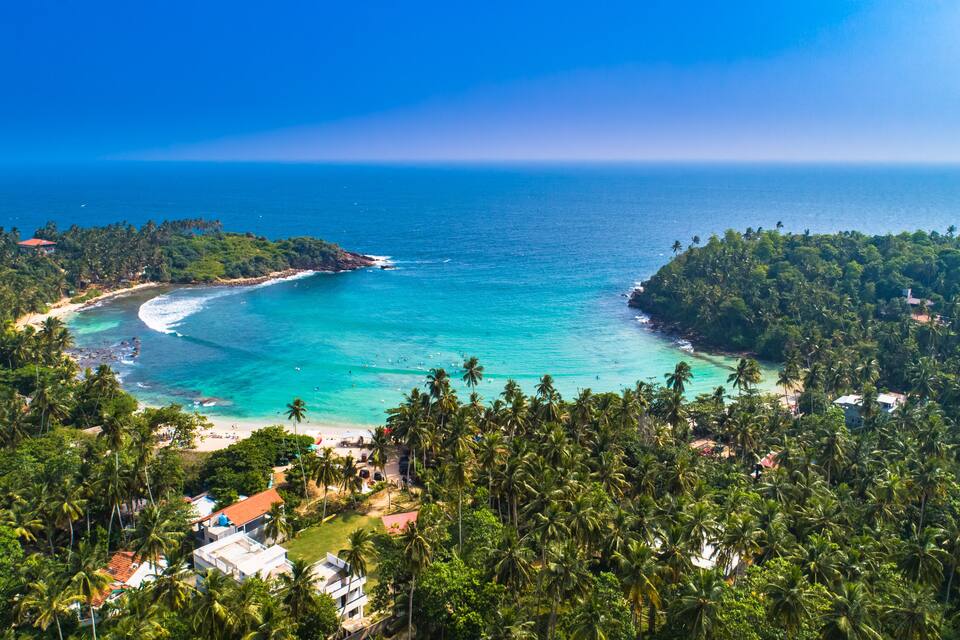 Ein tropischer Strand mit klarem, türkisblauem Wasser, umgeben von üppigen Palmen und einer kleinen Insel.