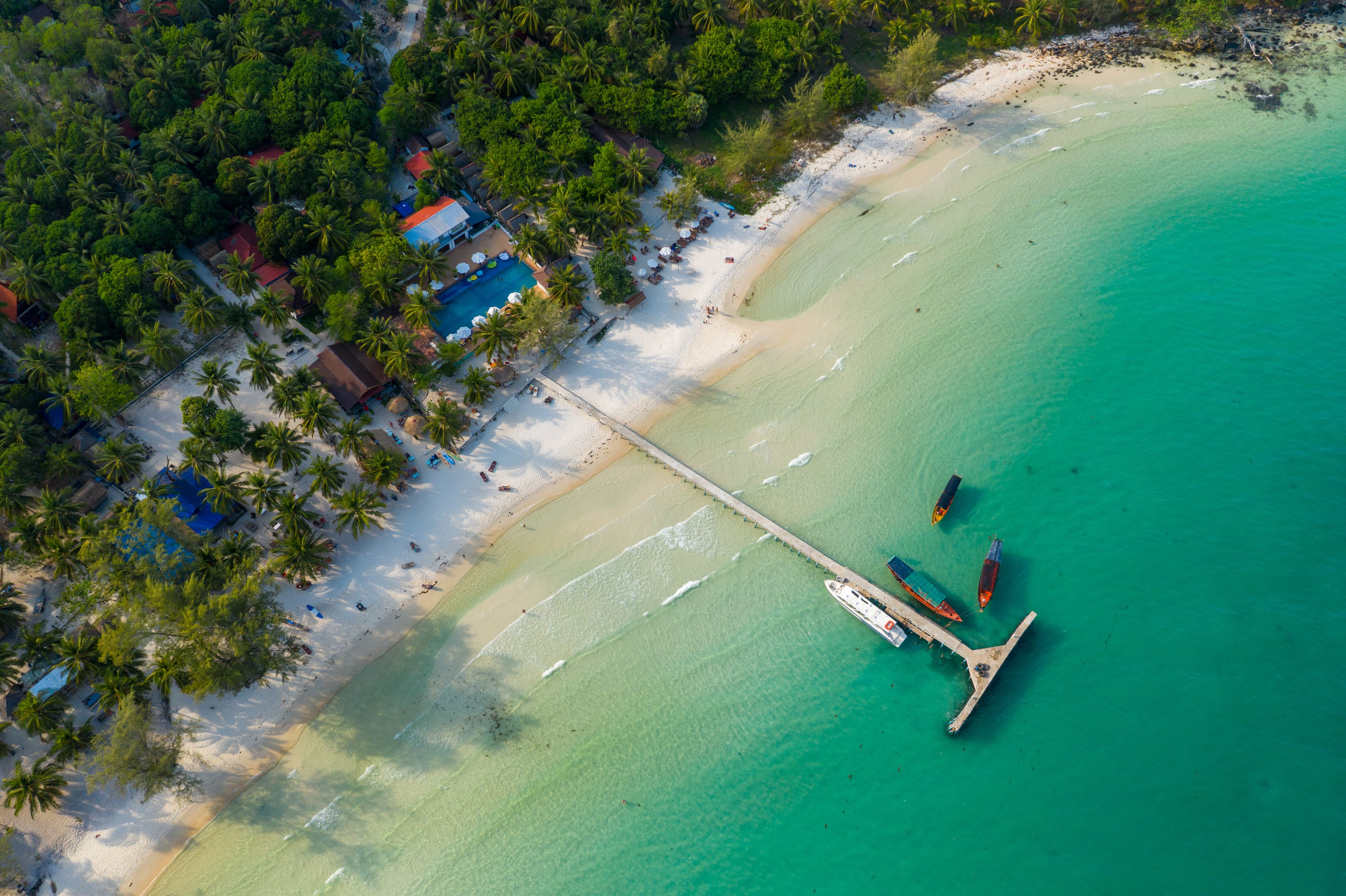 Overhead perspective shows a long wooden pier stretching from white sand into clear turquoise sea at Coconut Beach on Koh Rong island, Cambodia. Palm trees, scattered boats, and lush tropical