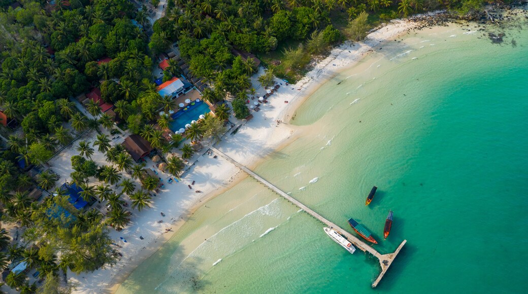 Overhead perspective shows a long wooden pier stretching from white sand into clear turquoise sea at Coconut Beach on Koh Rong island, Cambodia. Palm trees, scattered boats, and lush tropical