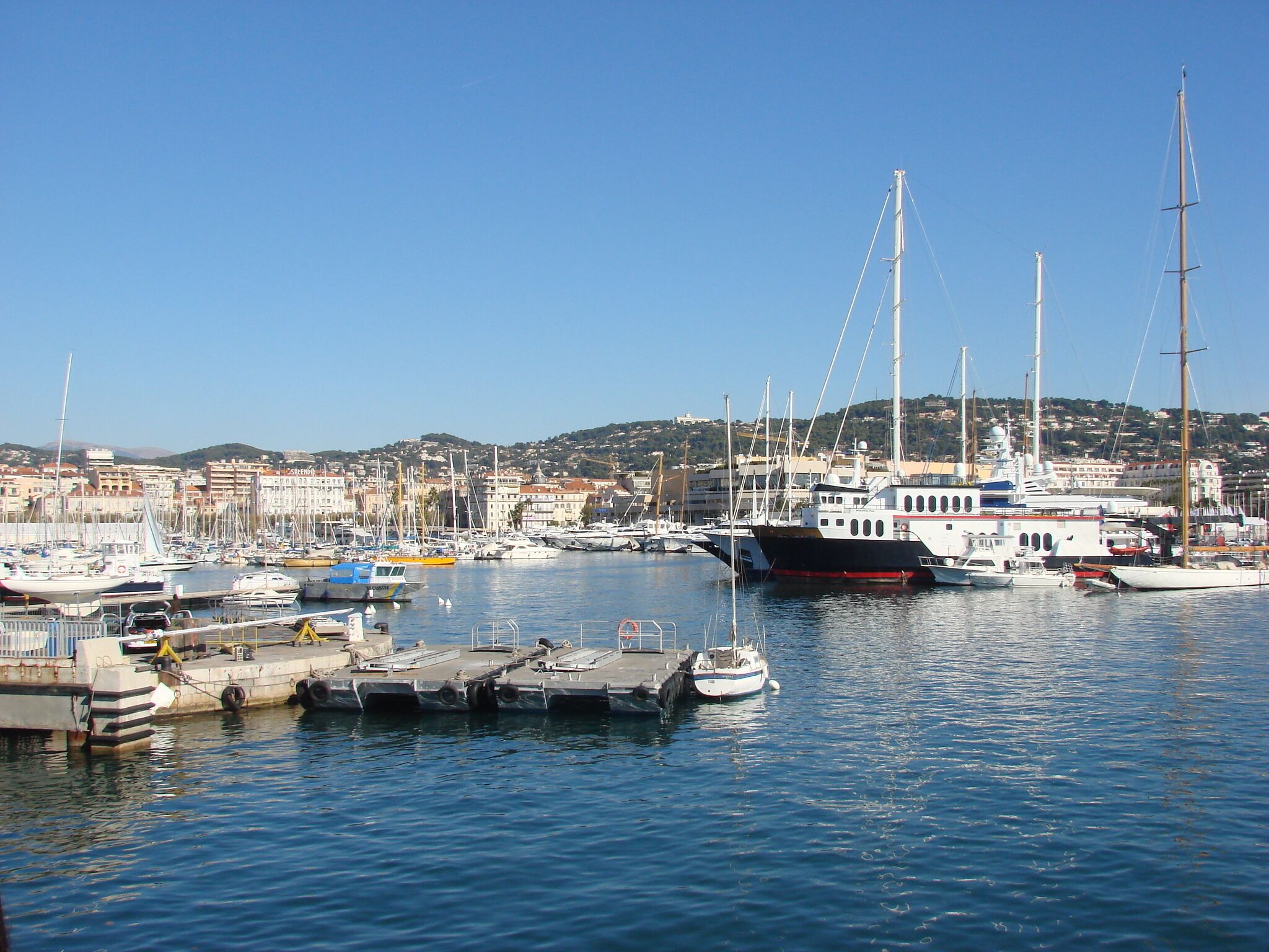A marina with various boats docked, a clear blue sky, and a distant town with buildings.
