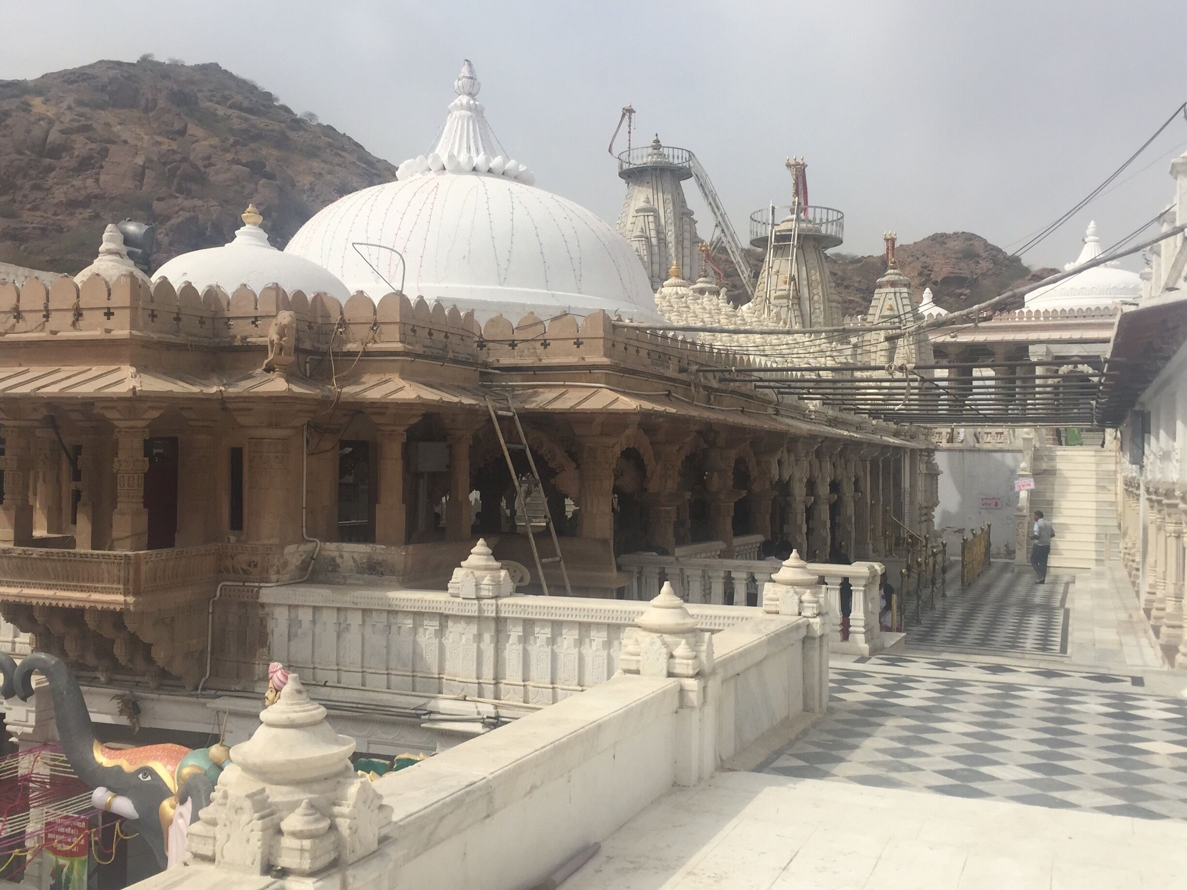 A temple with white domes and intricate carvings, a checkered pathway, and a mountainous backdrop.
